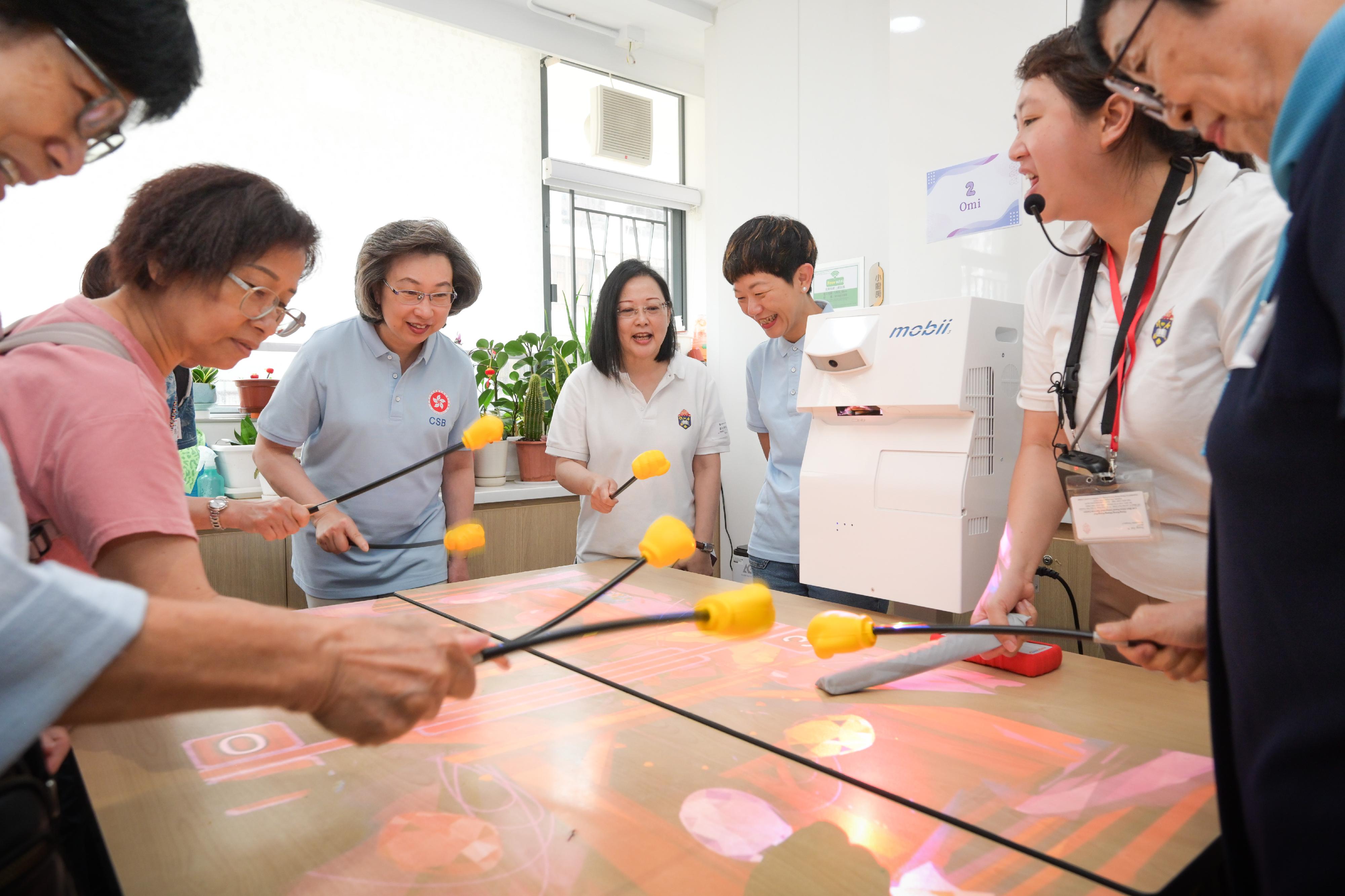 公務員事務局義工隊及數字政策辦公室義工小組今日（五月十七日）攜手到位於土瓜灣的香港聖公會樂民郭鳳軒綜合服務中心，進行「友智識」長者數碼共融計劃義工活動，輔助長者學習使用智能電話及應用程式，並一起互動體驗樂齡科技的樂趣，將科技融入日常生活之中。圖示（左三起）公務員事務局局長楊何蓓茵、香港聖公會福利協會總幹事劉冼靜儀、公務員事務局副秘書長張誼和公務員義工與長者一同參與感官統合訓練的遊戲，幫助提升長者的反應能力。
